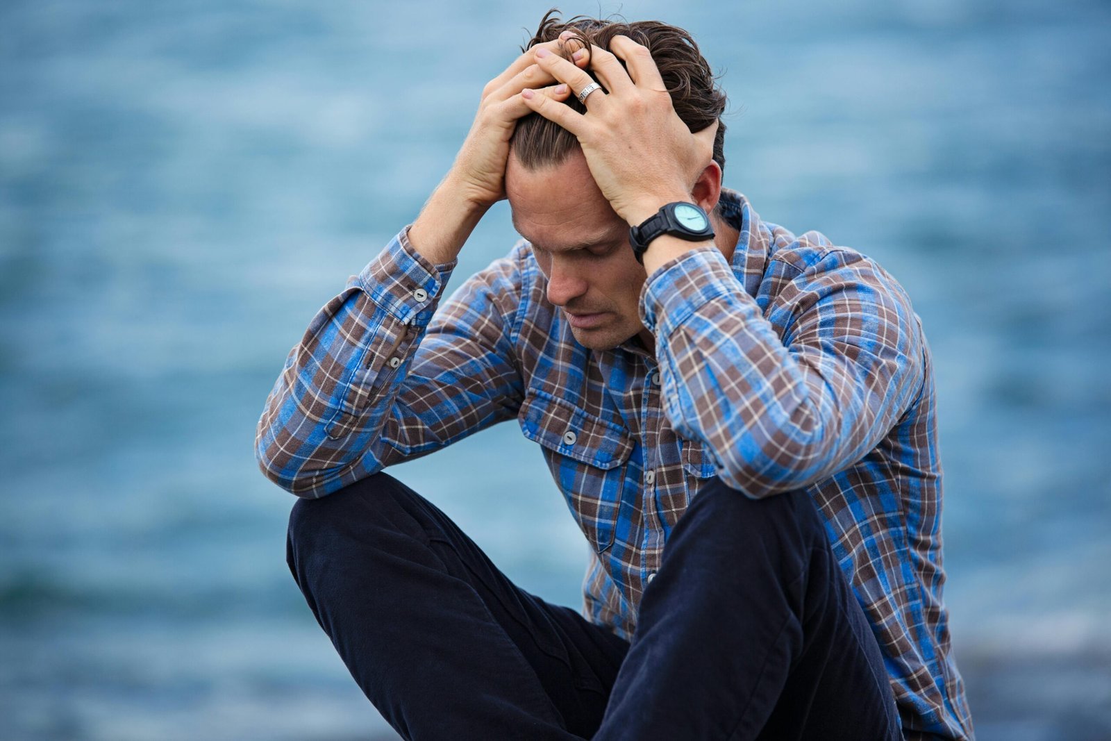 How To Cope With Anxiety Dissorder? 1 A man in a plaid shirt sits by the water looking distressed, symbolizing stress.